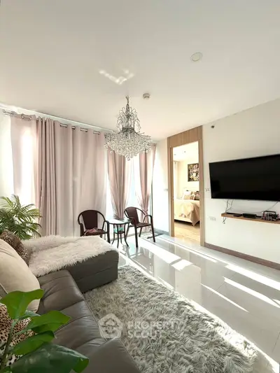 Elegant living room with chandelier, plush sofa, and wall-mounted TV, leading to a cozy bedroom.