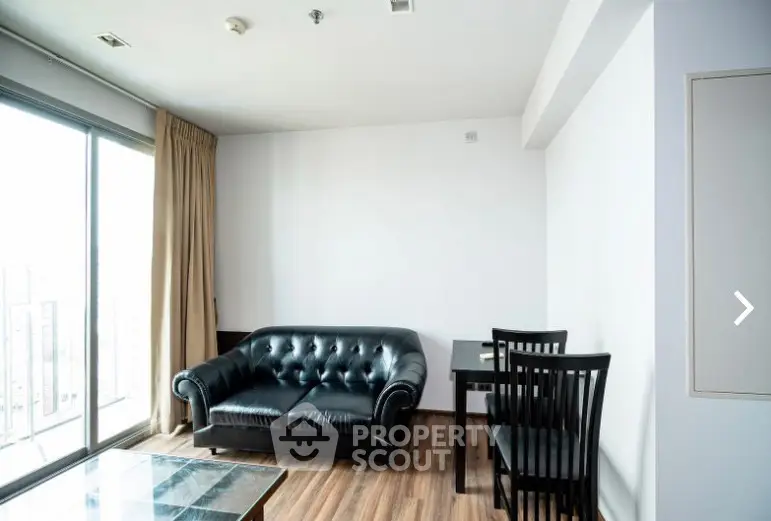 Modern living room with black leather sofa and dining set, featuring large windows for natural light.