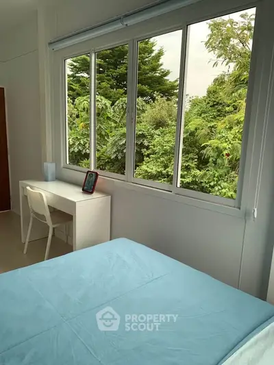 Bright bedroom with large window offering lush green views, featuring a cozy bed and a minimalist desk.