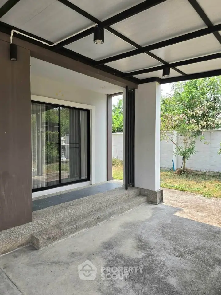 Modern home entrance with covered patio and sliding glass doors, surrounded by lush greenery.