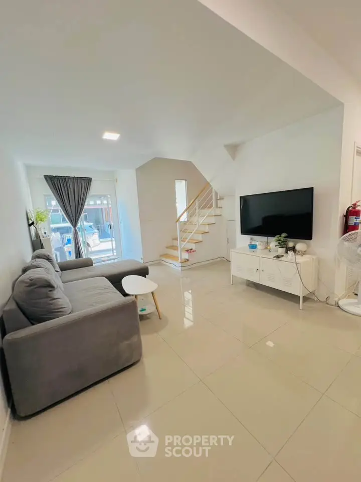 Modern living room with sleek furniture and staircase, featuring a large TV and natural light.