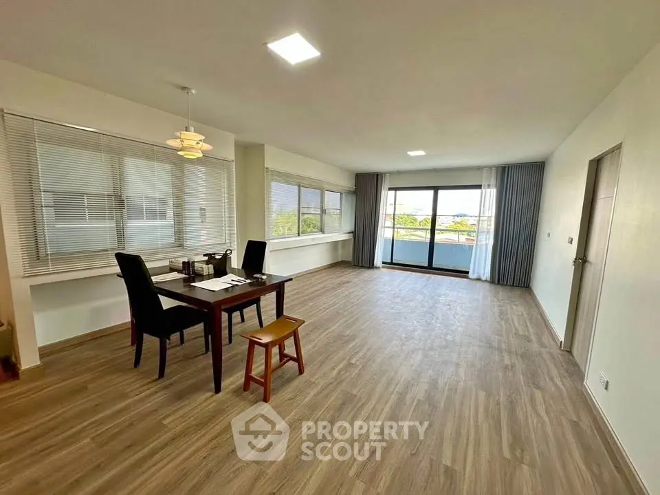 Spacious living room with large windows and balcony access, featuring modern dining setup and wood flooring.