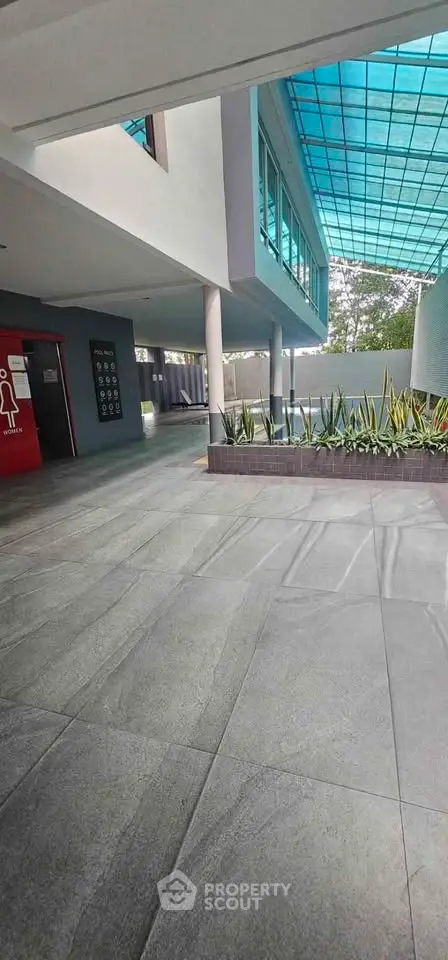Modern building entrance with sleek design and glass roof, featuring landscaped planters and spacious walkway.