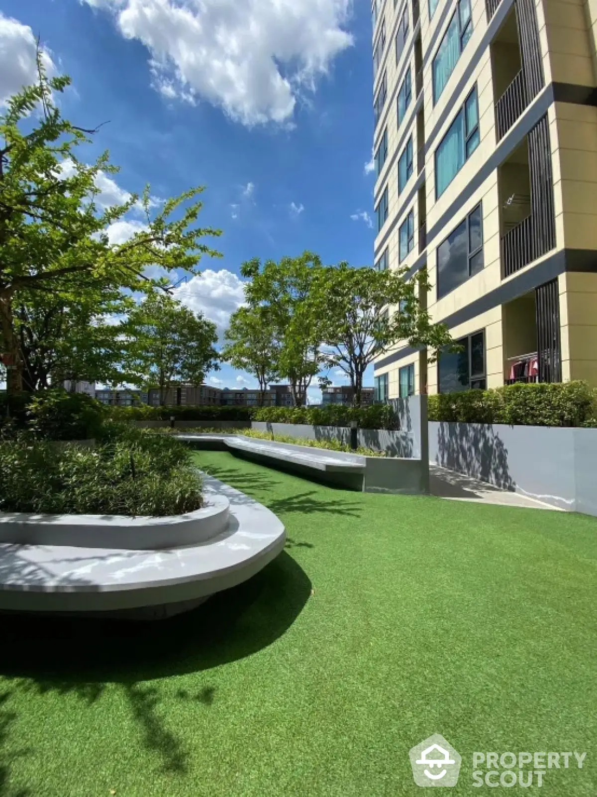 Modern apartment building with lush green garden and seating area under clear blue sky.