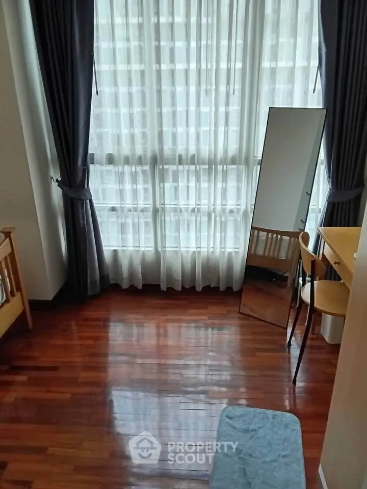 Bright bedroom with large window and wooden flooring, featuring a mirror and desk for a cozy living space.