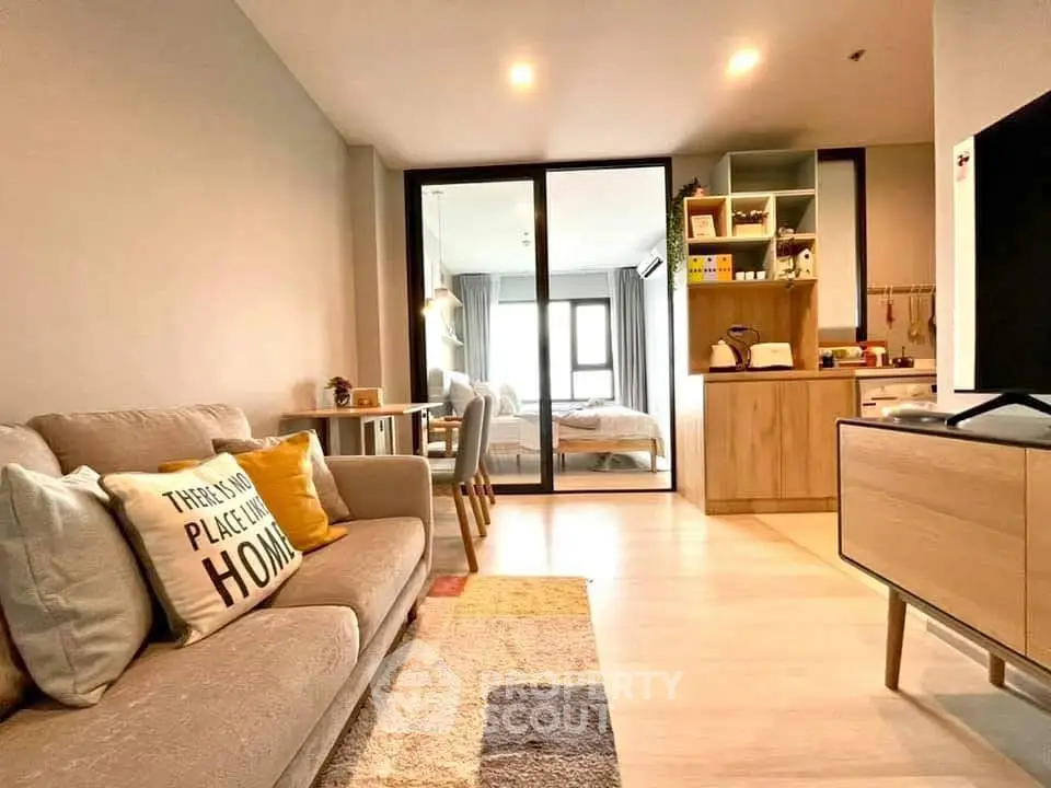 Modern open-plan living room with cozy sofa and stylish decor, leading to a bright bedroom through glass doors.