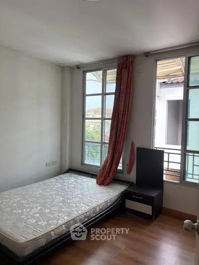 Cozy bedroom with large windows and natural light, featuring a simple bed and nightstand.