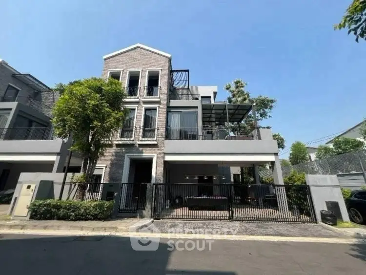 Modern three-story house with spacious balcony and gated entrance in sunny neighborhood.
