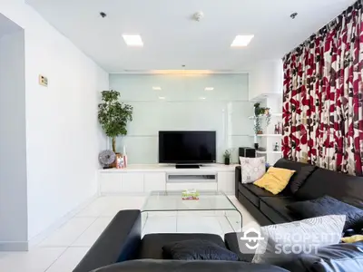 Modern living room with sleek black sofas, glass coffee table, and large TV, accented by vibrant curtains and greenery.