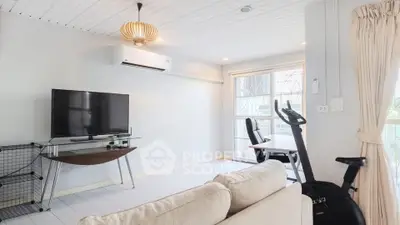 Bright living room with modern decor, featuring a TV, exercise bike, and natural light from large windows.