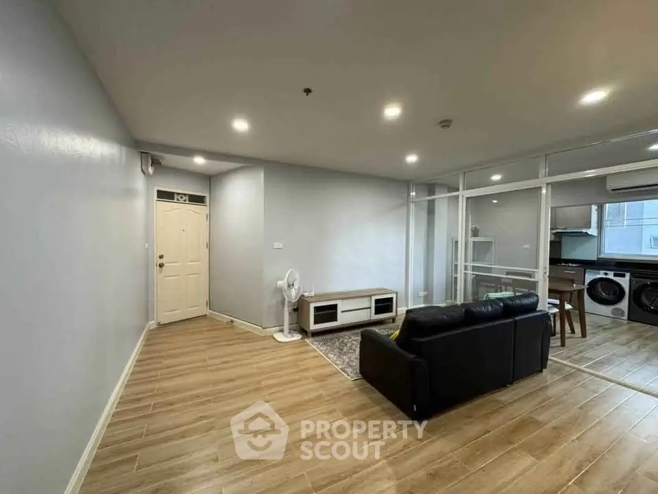 Modern living room with sleek wooden flooring and open kitchen layout, featuring a cozy black sofa and stylish decor.