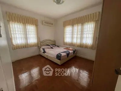 Cozy bedroom with parquet flooring and natural light from two windows, featuring a comfortable bed and elegant curtains.