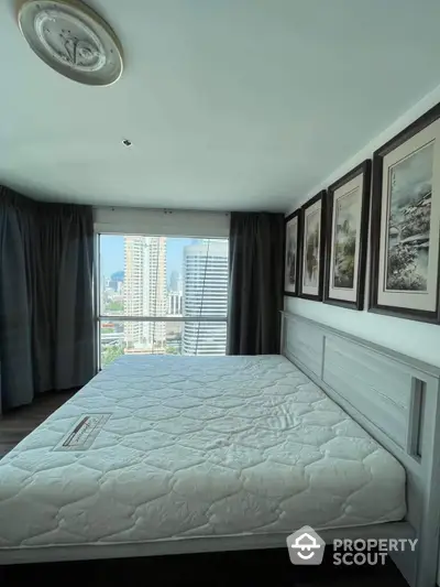 Spacious bedroom with large windows offering panoramic city views, complemented by elegant dark curtains and framed artwork, creating a serene urban retreat.