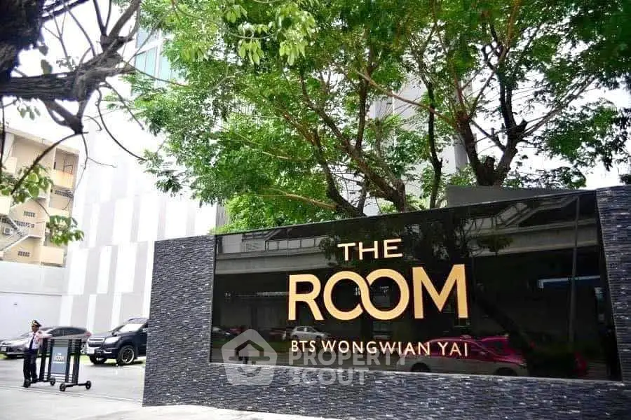 Modern building entrance with sleek signage at The Room BTS Wongwian Yai, surrounded by lush greenery.
