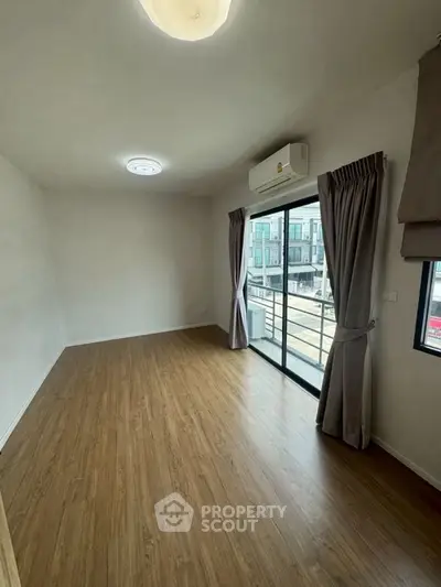 Spacious empty room with wooden flooring and large window offering natural light and balcony access.