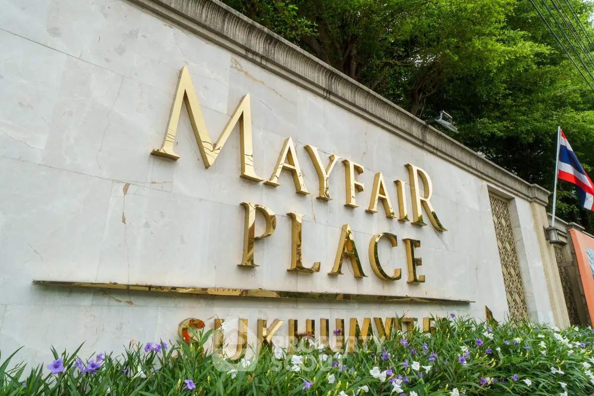 Elegant entrance sign of Mayfair Place Sukhumvit with lush greenery and flag, showcasing luxury real estate.