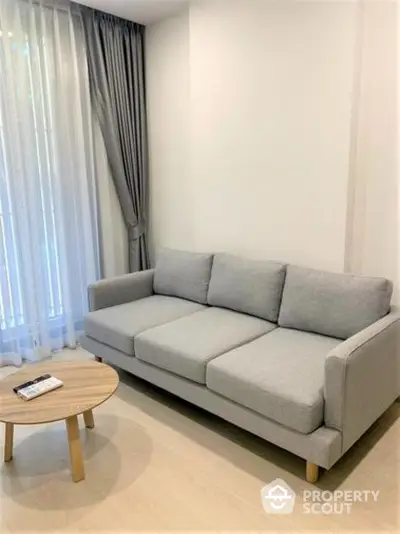 Modern minimalist living room with a comfortable grey sofa and a chic round wooden coffee table, complemented by sheer curtains for a cozy ambiance.