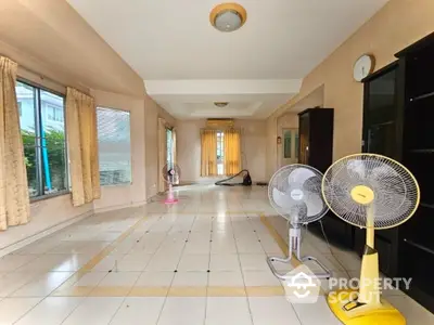 Spacious living room with large windows and tiled flooring, featuring standing fans and natural light.