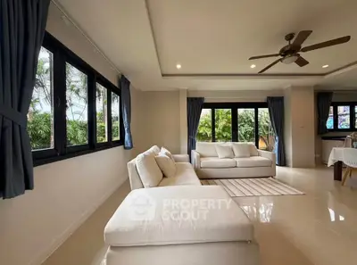 Spacious living room with large windows and garden view, featuring elegant white sofas and ceiling fan.