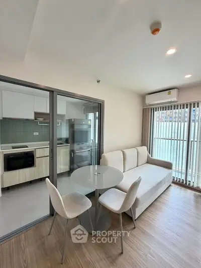 Modern living room with open kitchen and balcony view, featuring sleek furniture and natural light.