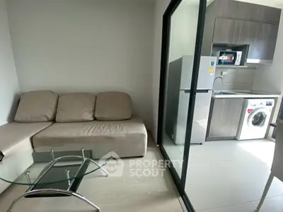 Modern compact living room with glass table and adjacent kitchen featuring washing machine and fridge.