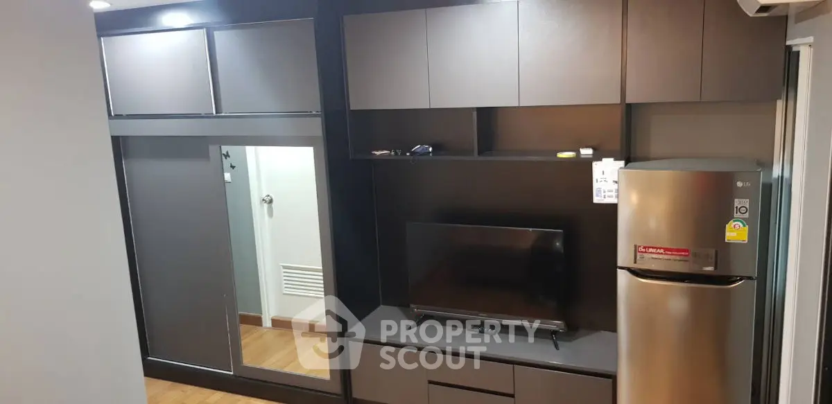 Modern living room with sleek cabinetry, TV, and stainless steel fridge in compact apartment.