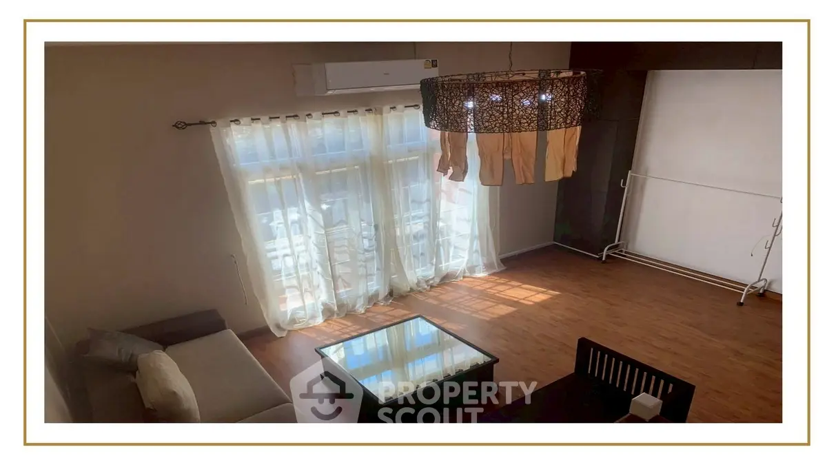 Spacious living room with large windows and elegant chandelier, featuring hardwood floors and modern furniture.