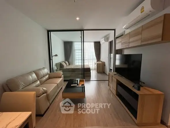 Modern living room with glass partition leading to bedroom, featuring sleek furniture and natural light.