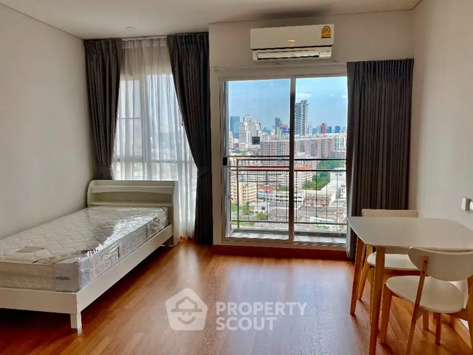 Modern bedroom with city view, featuring a single bed, dining table, and large window with balcony access.