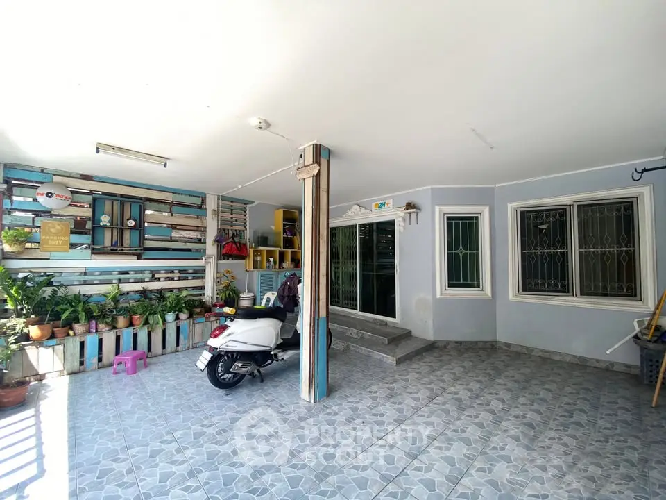 Charming home entrance with tiled flooring and scooter parking, surrounded by lush potted plants.
