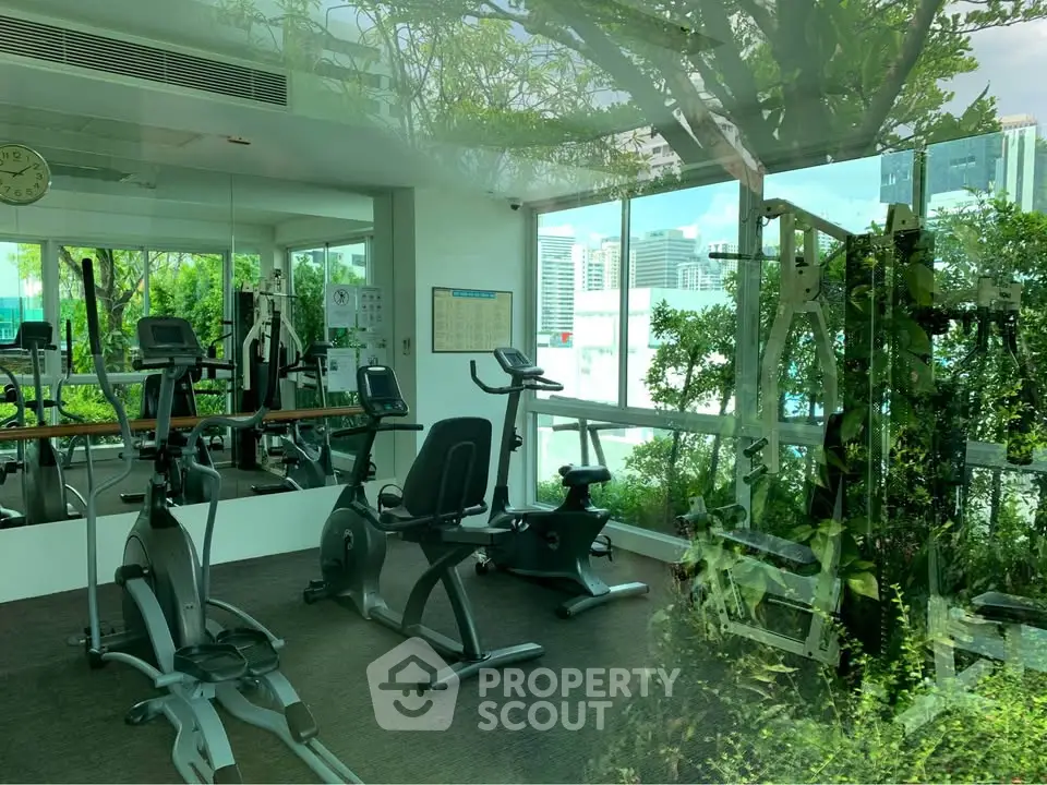 Modern gym with exercise equipment and city view, surrounded by greenery.