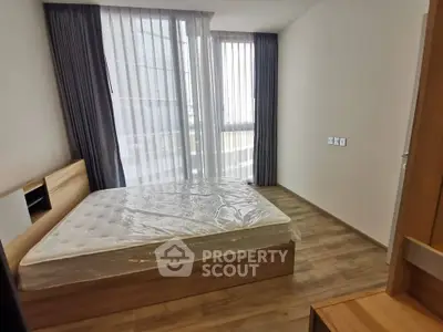 Modern bedroom with large windows and wooden flooring, featuring a new mattress and stylish curtains.
