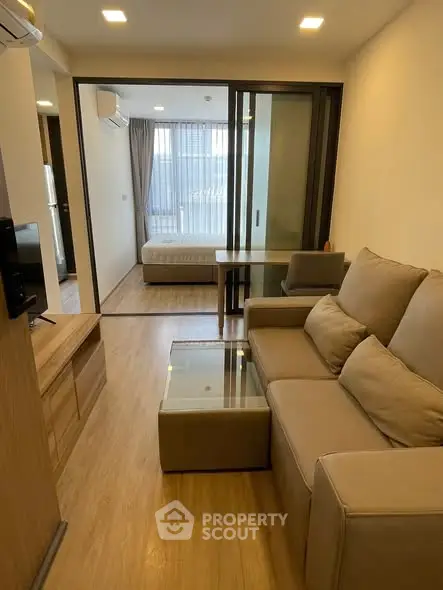 Modern living room with beige sofa and glass coffee table leading to a cozy bedroom.