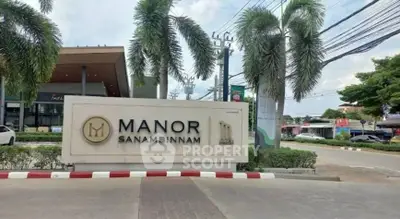 Entrance sign of Manor Sanambinnam with palm trees and street view.