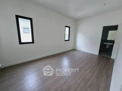 Spacious empty room with wooden flooring and two windows, leading to a small bathroom.