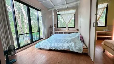 Bright bedroom with large windows and serene forest view, featuring minimalist decor and wooden flooring.