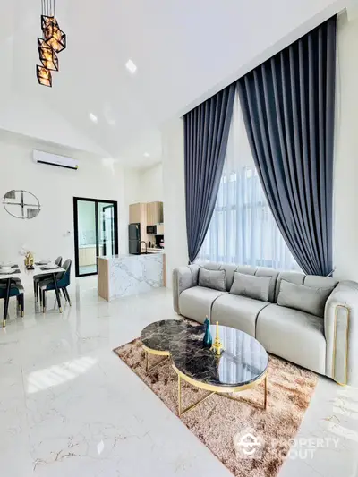 Luxurious open-plan living space with sleek marble flooring, plush grey sectional sofa, and elegant gold-accented coffee table, bathed in natural light from floor-to-ceiling windows draped with dark curtains.