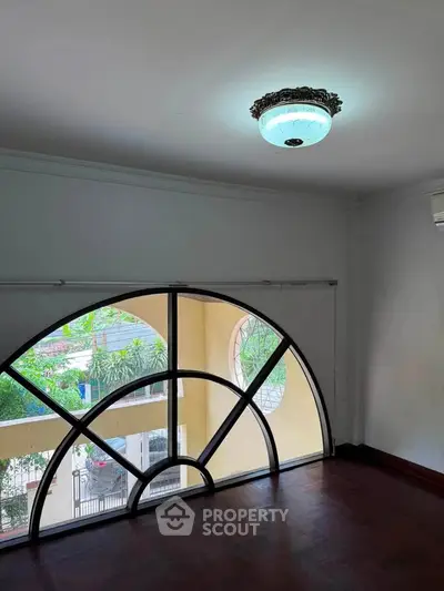 Spacious room with unique arched window and elegant ceiling light, offering a charming view of greenery.