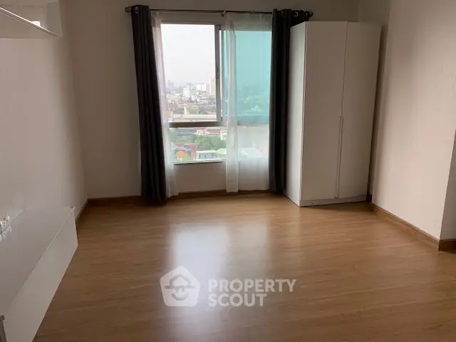Spacious empty room with wooden flooring and large window offering city view.