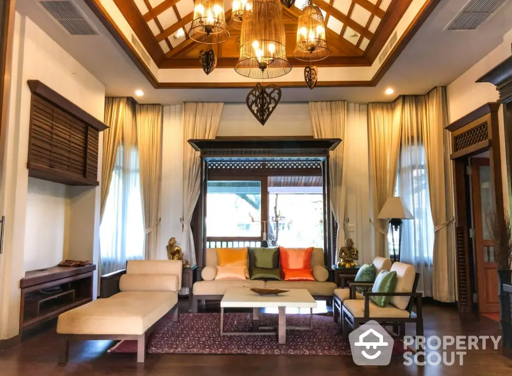 Luxurious living room with high ceiling, elegant chandelier, and plush seating, framed by rich wooden accents and warm lighting.