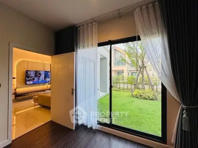 Modern living room with large window overlooking garden, elegant curtains, and open door to cozy interior.