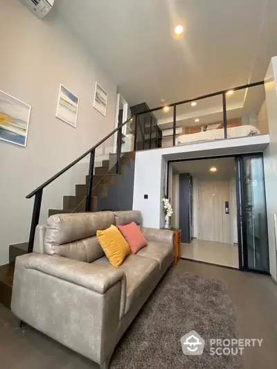 Modern duplex living room with plush grey sofa, elegant glass balustrade, and sleek staircase leading to an airy loft bedroom, accentuated by soft lighting and contemporary art.