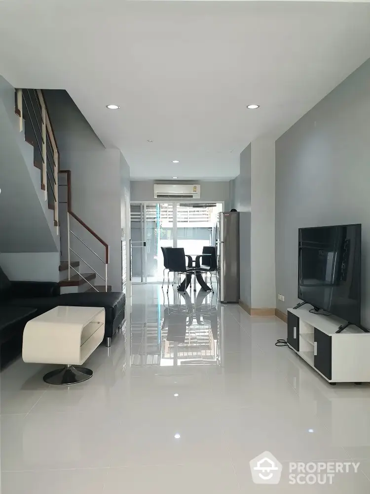 Modern living room with sleek furniture and open layout, featuring a staircase and dining area.