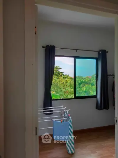 Serene bedroom with a large window offering a lush green view, complemented by dark curtains and wooden flooring, ideal for relaxation.