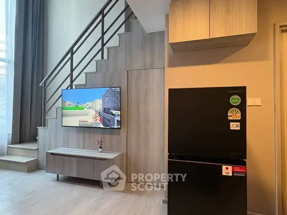 Modern living room with staircase, wall-mounted TV, and sleek black refrigerator.