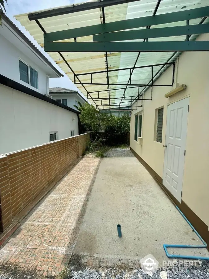 Spacious outdoor area with a protective overhead canopy and a brick wall, ideal for parking or outdoor activities, adjacent to a modern residential building.