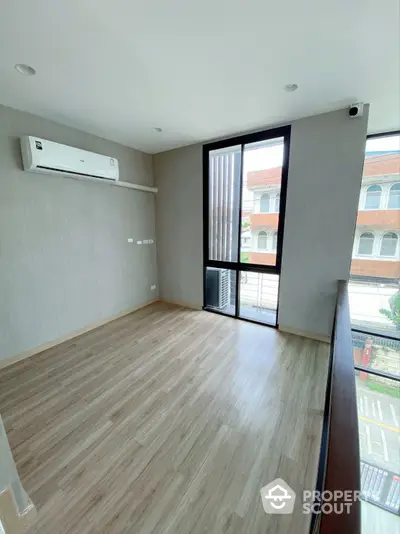 Spacious and well-lit empty room with polished wooden flooring, air conditioning unit, and a large window offering a view of the neighborhood.