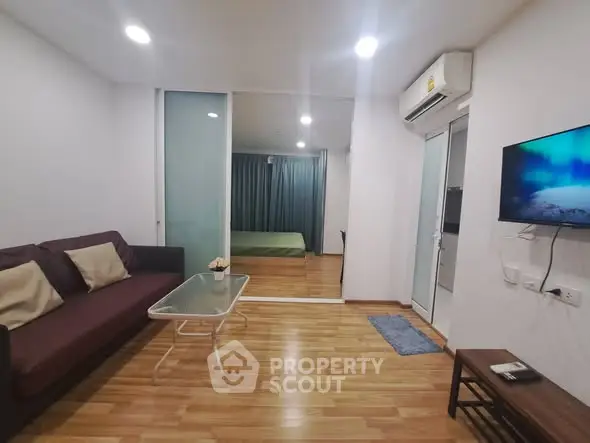 Modern living room with wooden flooring, cozy sofa, and wall-mounted TV, leading to a bedroom.