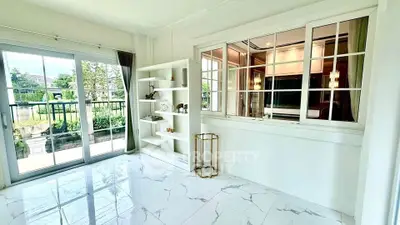 Bright living room with large windows and garden view, featuring elegant white shelving and marble flooring.