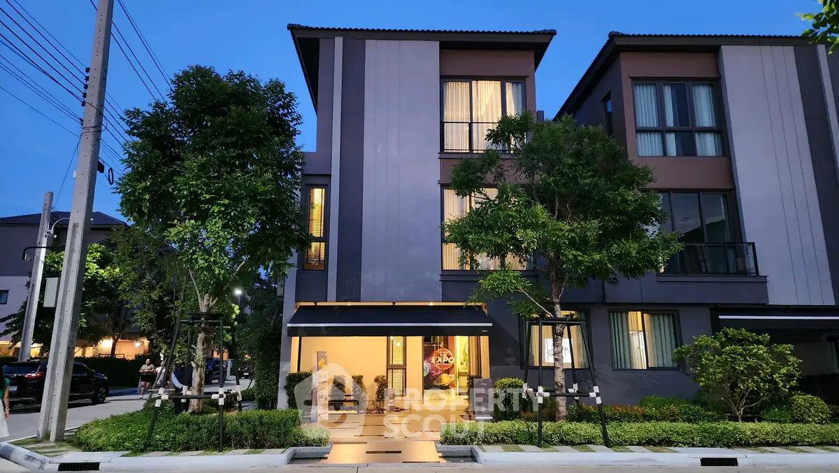 Modern three-story townhouse with illuminated entrance and lush greenery at dusk.